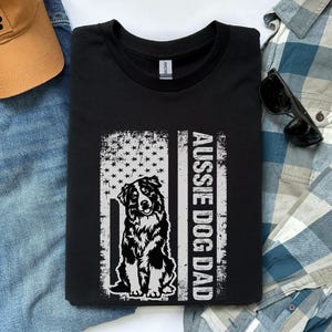 May include: Black t-shirt featuring a distressed American flag design with an Australian Shepherd dog illustration and the text "Aussie Dog Dad". The shirt is folded on a blue denim surface.