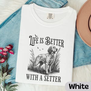 May include: White t-shirt with the text "Life is Better With A Setter" and a graphic of a setter dog in tall grass. The shirt is a Comfort Colors brand and is folded on top of blue jeans.