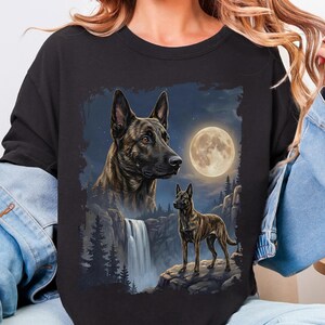 May include: Black sweatshirt featuring a detailed illustration of a dog with a waterfall, full moon, and forest scene. The dog is brown and black with a focused expression. The background includes a night sky with stars.