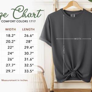 May include: A dark gray t-shirt displayed on a wooden hanger, with a size chart showing measurements in inches. The chart includes sizes S to 4XL, with corresponding width and length measurements. The text "Size Chart" is at the top.