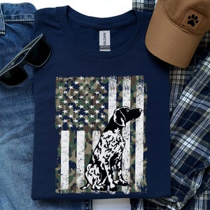 May include: A navy blue t-shirt with a distressed American flag design featuring a camouflage pattern and a black and white silhouette of a German Shorthaired Pointer dog.