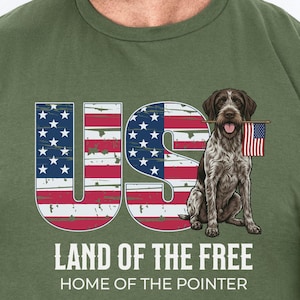 May include: Olive green t-shirt featuring a patriotic design. The graphic includes the letters "US" filled with the American flag, a dog holding a small flag, and the text "LAND OF THE FREE HOME OF THE POINTER".