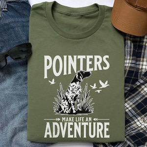 May include: Olive green t-shirt with the text "POINTERS MAKE LIFE AN ADVENTURE". The shirt features a black and white dog illustration, with three flying birds. Sunglasses, a brown cap, and blue jeans are also visible.