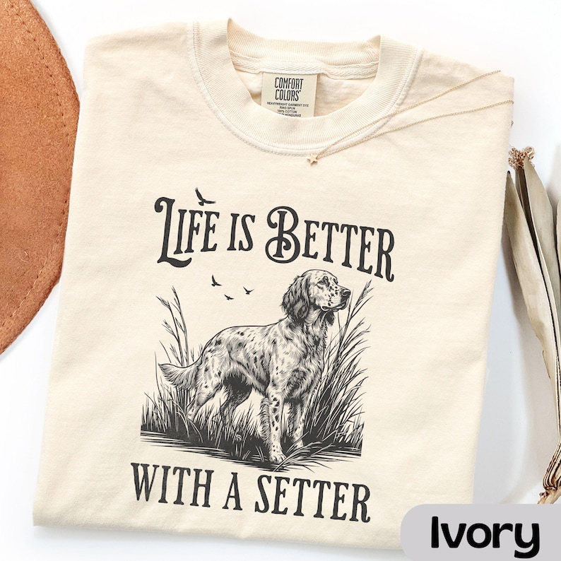 May include: Ivory-colored t-shirt with the text "Life is Better With A Setter" and a black and white illustration of a setter dog in tall grass. The shirt is a Comfort Colors brand.