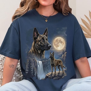 May include: Navy blue t-shirt featuring a detailed illustration of two dogs with a waterfall, full moon, and forest scene. The dogs are brown and black. The background is a night sky.