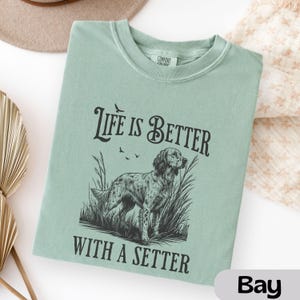 May include: A light green t-shirt with the text "Life is Better With a Setter" and a graphic of a setter dog in tall grass. The shirt is a crew neck style.