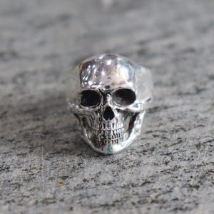 May include: A silver skull ring with detailed facial features, including eye sockets, a nose cavity, and teeth. The ring is set against a textured gray surface, showcasing the intricate design of the skull.
