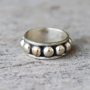 May include: A silver ring featuring a band of evenly spaced, raised, circular studs. The ring has a simple, classic design, and the metal has a slightly aged appearance. The ring is shown on a grey surface.