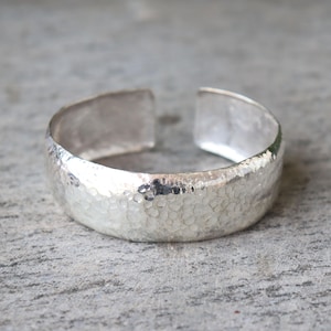May include: A silver cuff bracelet with a hammered texture. The open-ended design features a wide band with a textured surface, reflecting light. The bracelet is a simple, yet elegant piece of jewelry.