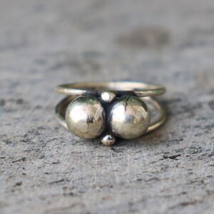 Puede incluir: Un anillo plateado con dos cuentas esféricas grandes y dos cuentas más pequeñas. El anillo tiene un diseño de doble banda, con las cuentas colocadas entre las bandas. El anillo está sobre una superficie gris texturizada.