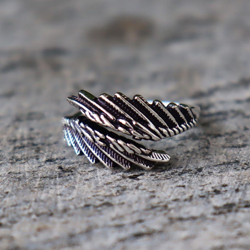 Puede incluir: Un anillo plateado con dise&ntilde;o de pluma. El anillo se envuelve alrededor del dedo con dos elementos de pluma detallados. Las plumas tienen l&iacute;neas y texturas intrincadas, creando una apariencia realista. El anillo est&aacute; sobre una superficie gris texturizada.