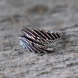 Puede incluir: Un anillo plateado con dise&ntilde;o de pluma. El anillo se envuelve alrededor del dedo con dos secciones de plumas detalladas. Las plumas tienen una apariencia texturizada, con l&iacute;neas y crestas individuales. El anillo est&aacute; sobre un fondo gris texturizado.