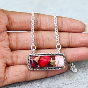 May include: A silver necklace with a rectangular pendant. The pendant features a mosaic of red, pink, and gold-colored stones. The chain is a delicate silver link. The pendant is approximately 1 inch wide.
