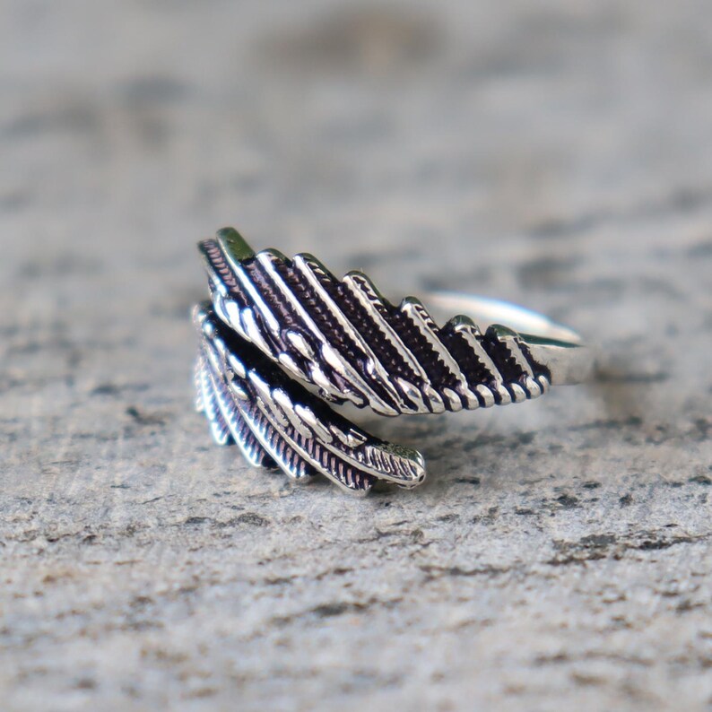 Puede incluir: Un anillo plateado con dise&ntilde;o de pluma. El anillo presenta patrones de plumas detallados, con las plumas curv&aacute;ndose alrededor del dedo. El anillo est&aacute; sobre un fondo neutro y texturizado.