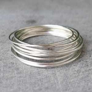 May include: A stack of silver-colored bangles, each a thin, smooth band. The bangles are arranged in a slightly offset circular pattern, creating a layered effect. The jewelry is displayed against a gray, textured surface.