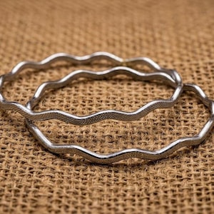 May include: Two silver-toned metal bangles featuring a continuous wavy, serpentine design. The surface of the bracelets has a fine, textured finish that catches the light, creating a subtle shimmer across the curved, undulating metal bands.