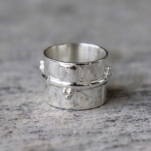 Könnte beinhalten: Ein silberfarbener Ring mit strukturierter Oberfläche und kleinen, erhabenen dekorativen Elementen. Der Ring hat ein breites Banddesign und eine polierte Oberfläche, geeignet für den täglichen Gebrauch.