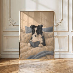 Op de afbeelding: Een ingelijste kunstprint met een zwart-witte Border Collie die rechtop in bed zit. De hond houdt een open blauw boek vast met zijn poten, omringd door beige kussens met patronen en een zachte blauwe deken in een gezellige slaapkamer.