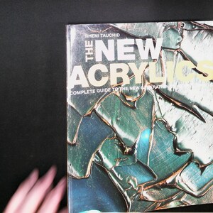 May include: A rectangular book titled The New Acrylics: Complete Guide to the New Generation of Acrylics by Rheni Tauchid. The cover features a textured, abstract design in shades of teal, green, and metallic gold with a glossy finish.