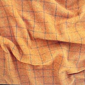 May include: Close-up of orange fabric with a black grid pattern. The fabric has a textured appearance and is draped, creating folds and shadows. The color is a warm, inviting orange.