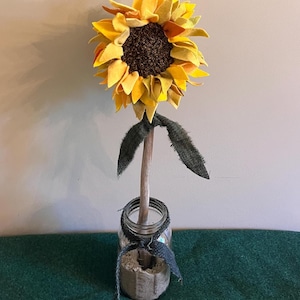 May include: A handcrafted sunflower made of yellow and orange felt petals with a dark brown textured center. The flower is attached to a light-colored wooden stem with two dark green fabric leaves, placed inside a clear glass jar filled with sand.