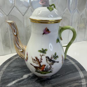 May include: A white porcelain teapot featuring a hand-painted scene of two birds perched on branches, surrounded by colorful butterflies. The lid is topped with a sculpted pink rosebud, and the handle and spout are accented with green and gold.