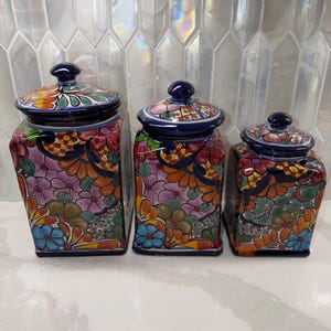 Könnte beinhalten: Ein Set aus drei quadratischen Keramikdosen mit passenden Deckeln, die jeweils mit einem lebendigen, handbemalten Blumenmuster in Orange, Blau, Pink und Grün auf dunkelblauem Grund verziert und nach Größe absteigend angeordnet sind.