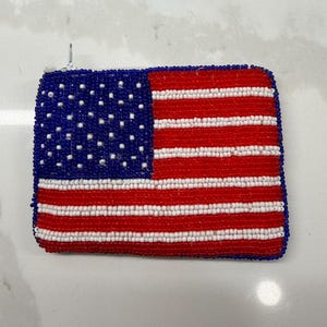 May include: A rectangular beaded coin purse featuring the American flag design. The purse has a blue field with white stars and red and white stripes. The purse is made of small, colorful beads.