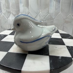 May include: A white ceramic bird figurine with blue accent lines. The bird is perched on a black and white checkered marble surface. The figurine has a glossy finish and a detailed design.
