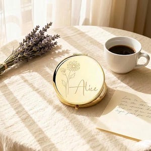 Könnte beinhalten: Ein goldener Kompaktspiegel mit floralem Design und dem eingravierten Namen "Alice". Eine Tasse Kaffee und ein Lavendelstrauß befinden sich ebenfalls auf dem Tisch. Eine handschriftliche Notiz ist ebenfalls vorhanden.
