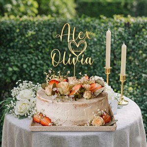 Op de afbeelding: Een versierde taart met een gouden taarttopper met de tekst "Alex Olivia 8.12.23". De taart is versierd met aardbeien, rozen en groen. Twee brandende kaarsen in gouden houders staan naast de taart.