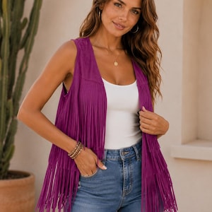 May include: A vibrant purple sleeveless waistcoat featuring two tiers of long fringe detailing that hangs from the chest and hemline. The garment is worn over a plain white vest top and blue denim jeans, showcasing a soft, suede-like texture throughout.