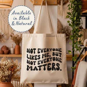 May include: A natural-colored canvas tote bag with two shoulder straps, featuring the black text "NOT EVERYONE LIKES ME, BUT NOT EVERYONE MATTERS." A circular graphic in the upper left corner states "Available in Black & Natural."