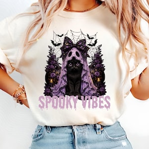 May include: A cream-colored t-shirt featuring a graphic of a black cat wearing a purple ghost-like hood with a large bow. The design includes spiderwebs, bats, dark roses, and the text SPOOKY VIBES in a light purple, dripping-style font.