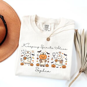 May include: A cream-colored short-sleeve t-shirt featuring a graphic with the text "Keeping Spirits Alive" and "Sophia". The design includes three cartoon characters dressed as medical staff, surrounded by pumpkins, bats, and stars.