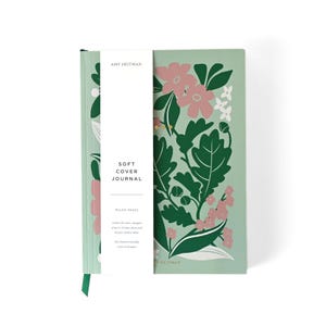 May include: A soft-cover journal featuring a floral pattern. The cover is a pale green with pink flowers and green foliage. A white band with the text "SOFT COVER JOURNAL" is wrapped around the journal. It includes a green ribbon bookmark.