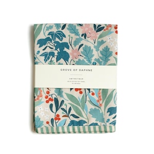 May include: A beige tea towel with a floral pattern in shades of blue, green, pink, and red. The towel has a green and white striped border and a label that reads "Grove of Daphne Amethystman 100% Cotton Tea Towel 20 x 28 inches".