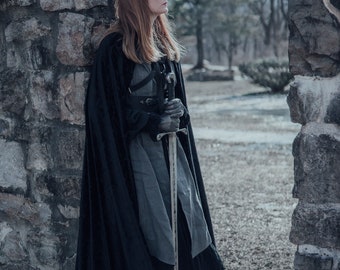 May include: A woman in a black cloak and grey dress stands in a stone doorway, holding a sword. She is looking off to the side, and the background is a blurry forest.
