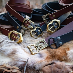 May include: A collection of leather belts with various metal buckles. The belts are brown and black and the buckles are gold and silver.