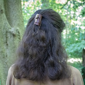 May include: A brown leather hair clip with a bone accent. The clip is being used to hold back a person's long, dark brown hair.