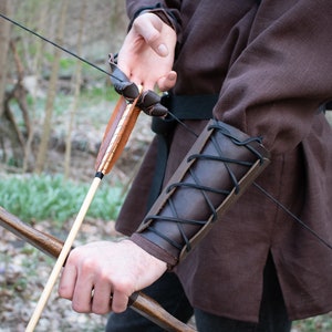 May include: A person wearing a brown tunic and leather arm guard holds a wooden bow and arrow. The arrow is fletched with brown feathers.