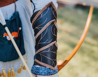 Op de afbeelding: Een persoon houdt een houten boog en pijl vast. De persoon draagt een bruine leren armbeschermer met een decoratief ontwerp. De armbeschermer is vastgemaakt met een blauw-witte band met een patroon.