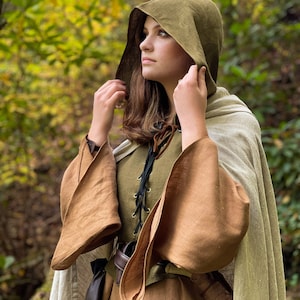 Pode incluir: Uma mulher vestindo um manto marrom com capuz, um corpete de linho verde e mangas marrons. O capuz está puxado sobre a cabeça e ela está olhando para cima para algo fora da câmera. O fundo é uma floresta desfocada.