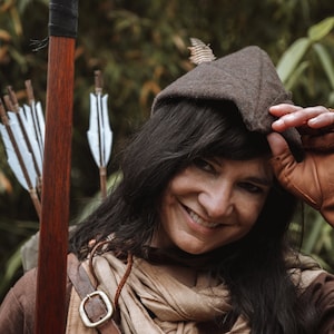 以下が含まれることがあります： 茶色のフード付きチュニックと真鍮のバックルが付いた茶色の革のベルトを着用した女性。彼女は左手に弓矢を持ち、カメラに向かって微笑んでいます。