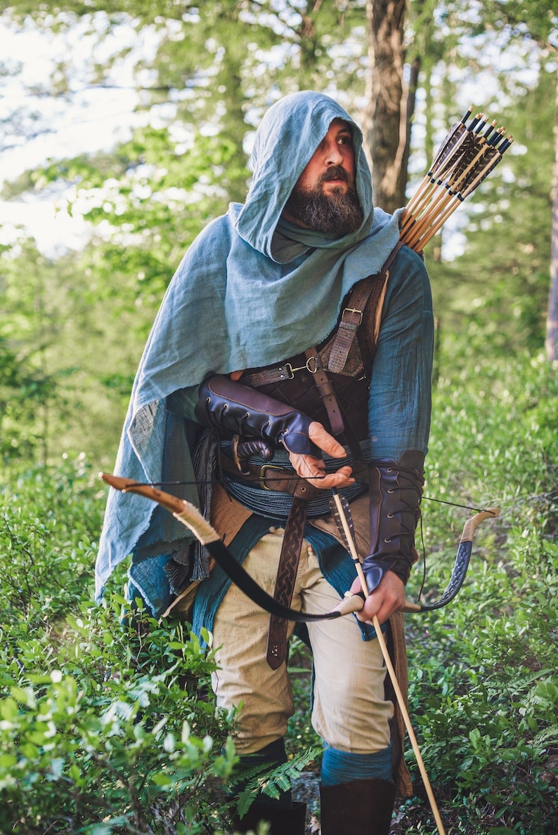 May include: A person wearing a blue hooded cloak and leather armor holds a bow and arrow in a forest setting.