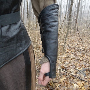 May include: Black leather arm guard with a strap and buckle closure. The guard is worn on the left arm and extends from the wrist to the elbow.