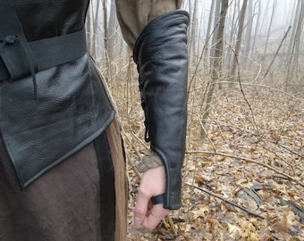 May include: Black leather arm guard with a strap and buckle closure. The guard is worn on the left arm and extends from the wrist to the elbow.