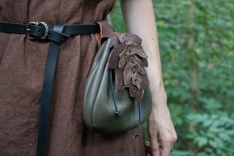 May include: A brown leather pouch with a drawstring closure and a decorative leaf design. The pouch is attached to a black leather belt.