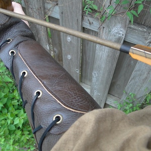 May include: A brown leather arm guard with black laces and brass eyelets. The arm guard is worn on the left arm and is secured with a strap.