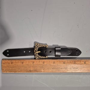 May include: A black leather belt with an ornate gold-colored buckle. The belt has a rectangular shape with a series of holes for adjustment. The buckle features a detailed, vintage design. The belt measures approximately 8 inches in length.